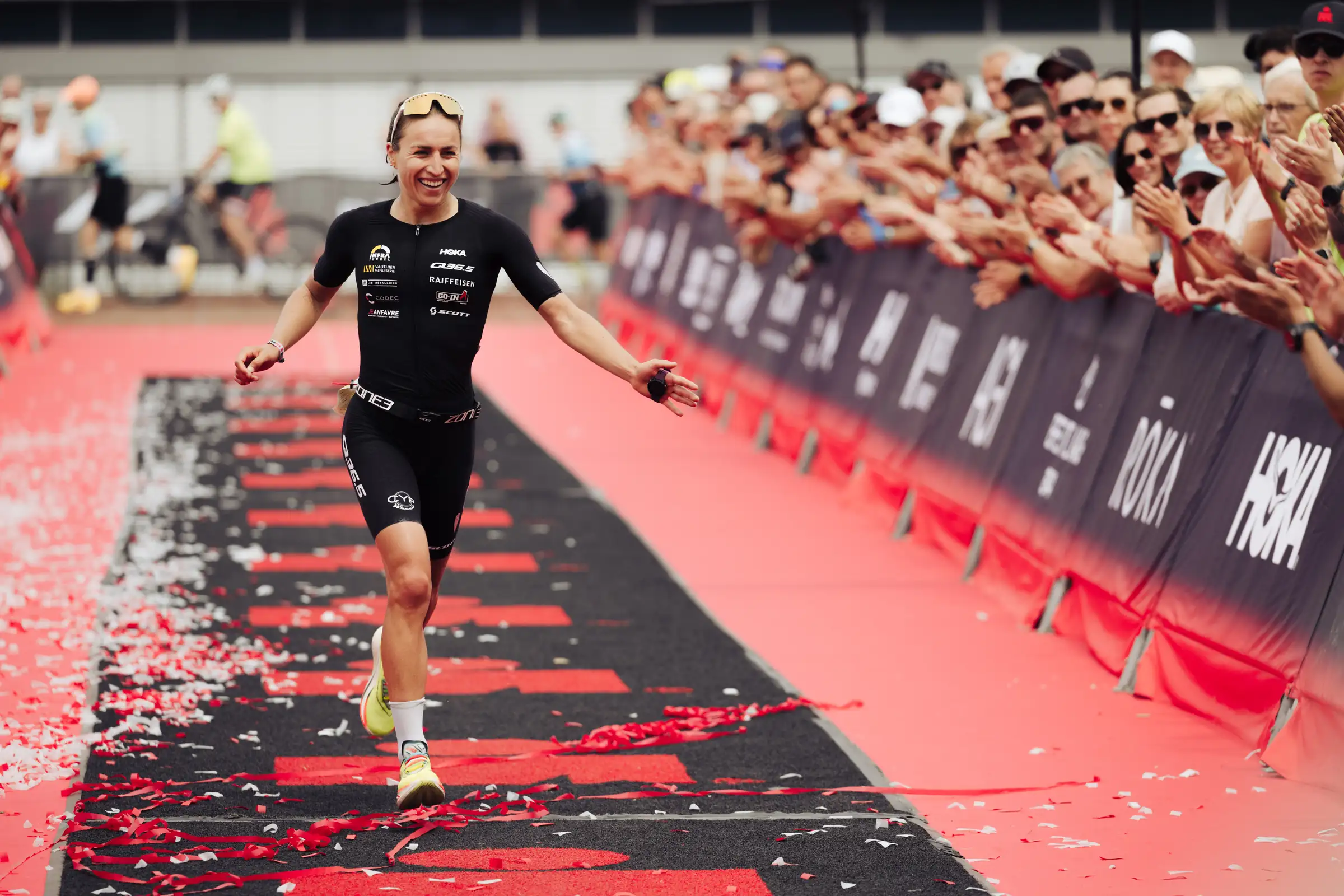 Loanne Duvoisin on the red carpet of an Ironman finish line, arms open, the crowd applauding
